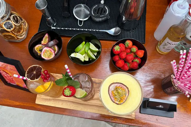 Bovenaanzicht van de bar met drie cocktails op een olijfhouten plank, bakjes met passievrucht, limoen en aardbeien ernaast en het barmateriaal erachter